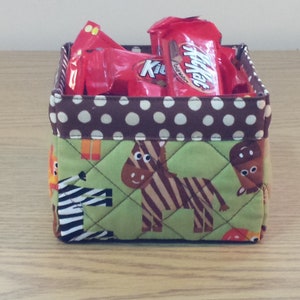 May include: A brown and white polka dot fabric storage bin with a green fabric bottom featuring a zebra and giraffe print. The bin is filled with red and white candy wrappers.
