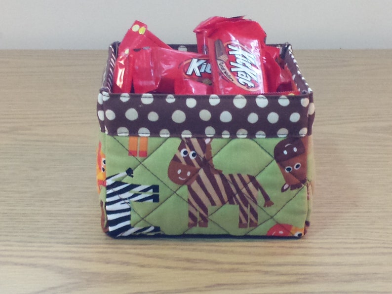 May include: A brown and white polka dot fabric storage bin with a green fabric bottom featuring a zebra and giraffe print. The bin is filled with red and white candy wrappers.