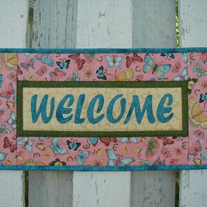 May include: A rectangular welcome sign with a pink background adorned with colorful butterflies. The sign features the word "WELCOME" in teal script within a beige and green border, framed by a teal border. The sign is attached to a white picket fence.