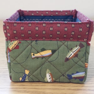 May include: A small, square fabric basket with a quilted design. The exterior features a green fabric with fish illustrations, while the top border is a maroon pattern. The interior lining is a blue and white pattern.