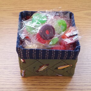 May include: A small, square fabric basket filled with wrapped candies. The basket has a green quilted exterior with a fish pattern and a blue patterned trim. Candies are various colors, including red, green, and purple.
