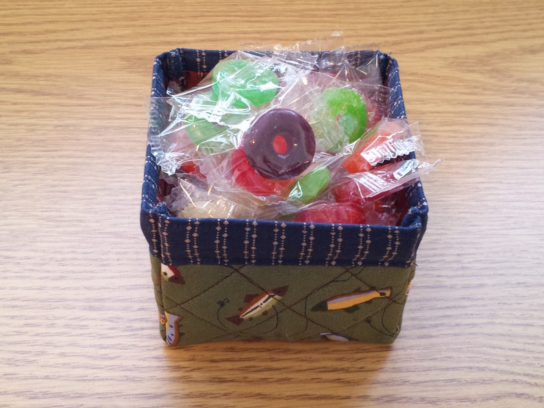 May include: A small, square fabric basket filled with wrapped candies. The basket has a green quilted exterior with a fish pattern and a blue patterned trim. Candies are various colors, including red, green, and purple.