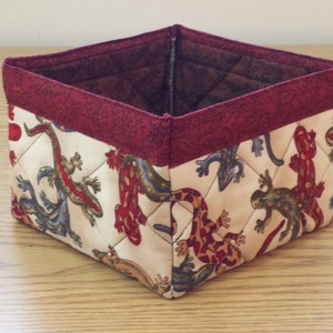 May include: A square-shaped fabric storage basket with a red trim. The basket is decorated with a pattern of colorful lizards on a cream background.