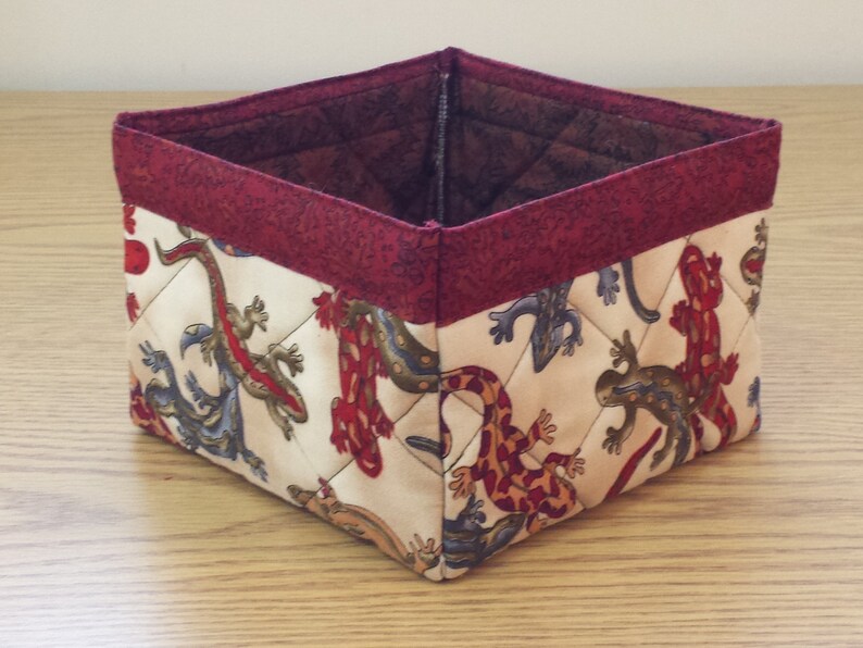 May include: A square-shaped fabric storage basket with a red trim. The basket is decorated with a pattern of colorful lizards on a cream background.