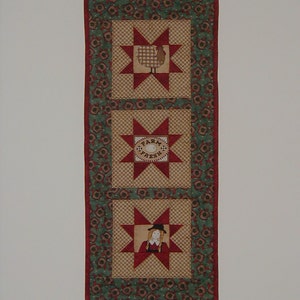 May include: A red, green, and brown quilted table runner with three star-shaped patches. The top patch features a brown and white checkered chicken, the middle patch has a white circle with the text "Farm Fresh", and the bottom patch has a girl wearing a red dress and a black hat.