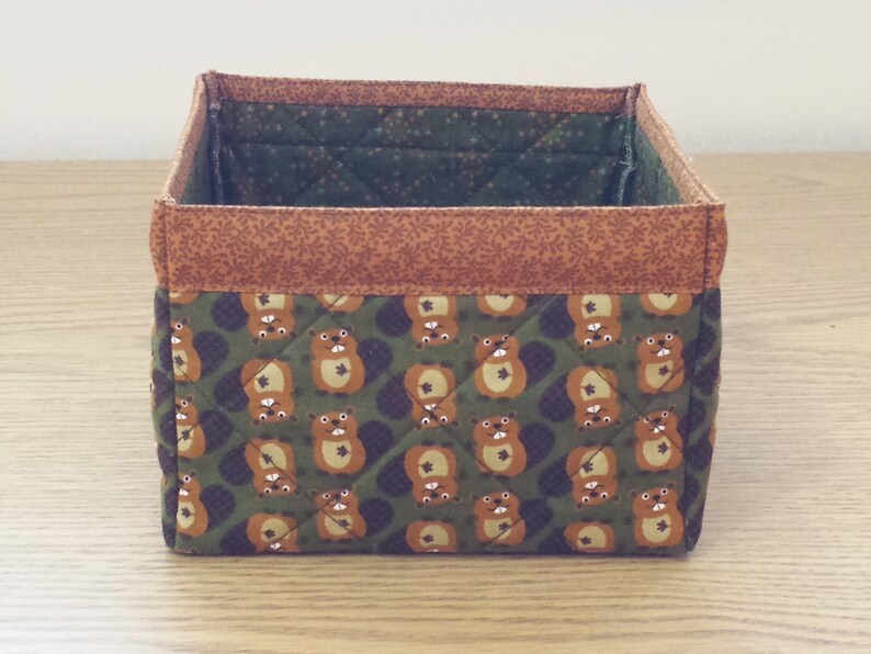 May include: A brown and green fabric storage box with a quilted pattern of cartoon beavers. The box has a brown fabric trim around the top.