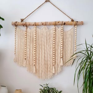Pode incluir: Uma tapeçaria de macramé bege com contas de madeira e borlas. A tapeçaria está presa a um galho natural.