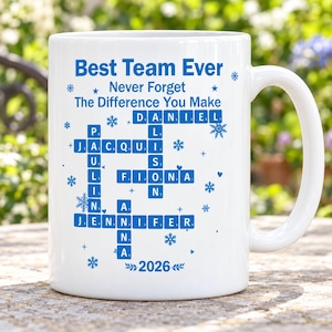 May include: A white ceramic mug featuring blue text and a crossword-style grid of names including Daniel, Jacqui, Alison, Fiona, Paulina, Jennifer, and Anna. The design includes the text Best Team Ever, Never Forget The Difference You Make, and 2026.