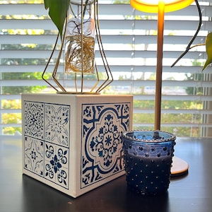 May include: A square white box featuring blue tile-style patterns on its sides, topped with a gold geometric wire frame holding a glass vase with plant roots. Beside it sits a small, dark blue glass candle holder with a textured hobnail design.
