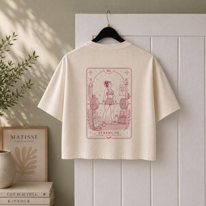 May include: A cream-coloured short-sleeve t-shirt featuring a maroon graphic on the back. The design depicts a person lifting a barbell in a gym, framed like a tarot card with the text VIII, STRENGTH, and COURAGE, FOCUS, RESILIENCE printed below.