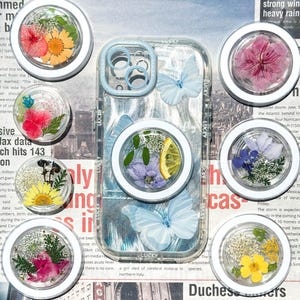 Puede incluir: Una funda transparente para teléfono con un marco azul claro y detalles de mariposas, rodeada de varios pop sockets circulares blancos. Cada pop socket contiene flores secas y una rodaja de limón, mostrando un tema floral.
