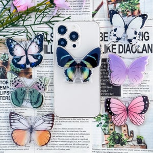 Peut inclure: Une collection de supports de téléphone en forme de papillon aux couleurs vives. Les supports présentent divers motifs, notamment des papillons roses, violets, bleus et blancs. Ils sont présentés sur un fond de journal avec des accents floraux.