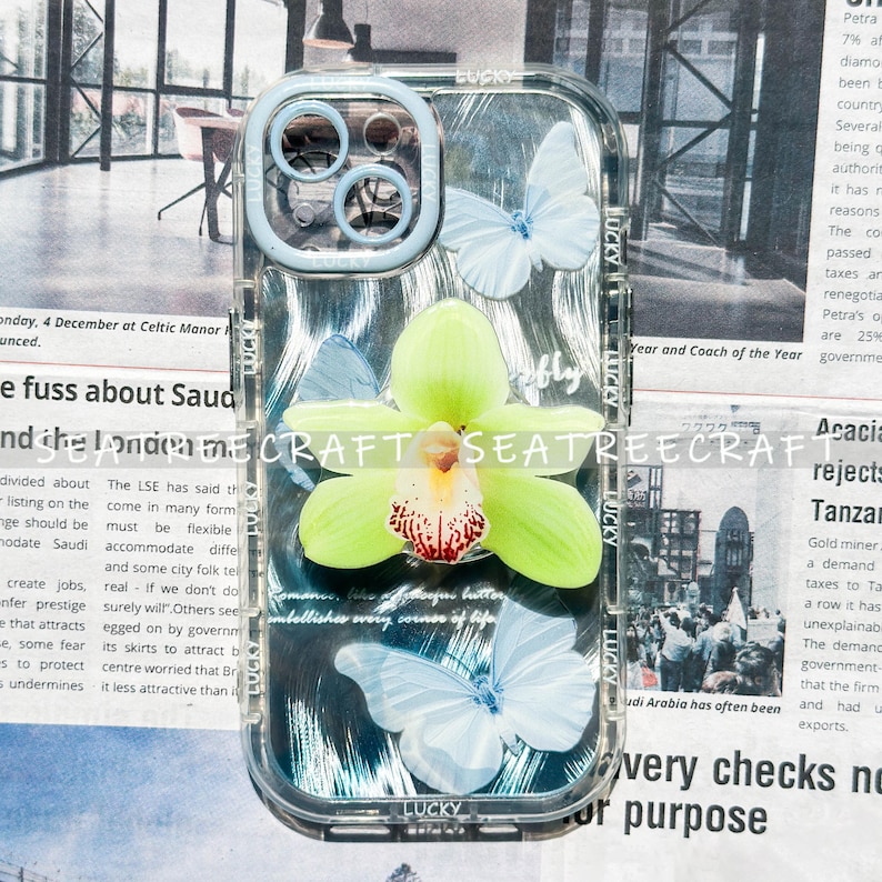 May include: Clear phone case with a light blue bumper, featuring a large green orchid and three white butterflies. The case has the word "Lucky" printed on it. The background is a newspaper.