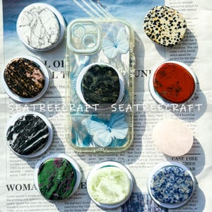 Könnte beinhalten: Eine Auswahl an Handyhaltern mit verschiedenen Designs, darunter Marmor und Sprenkelmuster, sowie eine transparente Handyhülle mit Schmetterlingsmotiven. Die Halterungen sind rund und auf weißen Basen befestigt.