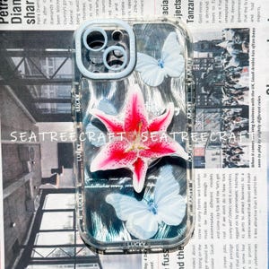 May include: Clear phone case with a floral and butterfly design. The case features a large pink and white lily and three light blue butterflies. The text "SEATREECRAFT" is visible on the case.