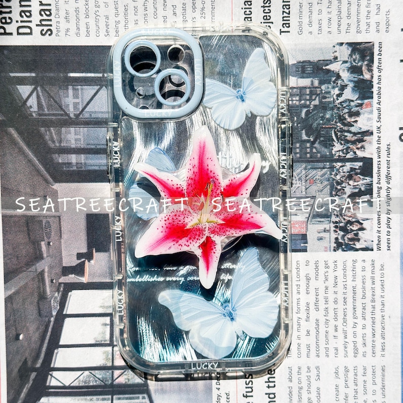 May include: Clear phone case with a floral and butterfly design. The case features a large pink and white lily and three light blue butterflies. The text "SEATREECRAFT" is visible on the case.