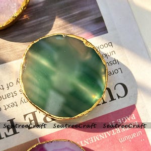Pode incluir: Uma fatia circular de ágata verde com uma superfície polida e translúcida e uma borda texturizada de cor dourada metálica galvanizada. A pedra apresenta variações naturais na profundidade da cor e na transmissão da luz, criando uma aparência lisa.