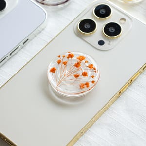 Peut inclure: Un support de téléphone transparent avec un motif de fleurs orange séchées. Le support rond est fixé à un téléphone blanc avec un cadre doré. Le support présente un design transparent avec des détails floraux.