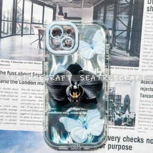 May include: Clear phone case with a black orchid and white butterflies design. The case has a light blue border and the word "LUCKY" is printed on the case. The background is a newspaper print.