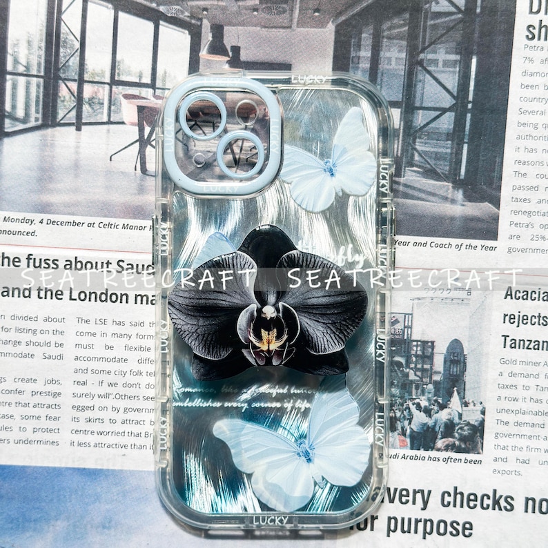 May include: Clear phone case with a black orchid and white butterflies design. The case has a light blue border and the word "LUCKY" is printed on the case. The background is a newspaper print.