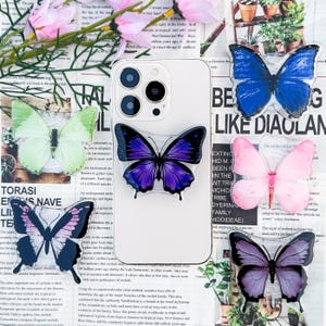 May include: A white iPhone case with a clear back, decorated with colorful butterfly stickers. The stickers are in various shades of purple, blue, green, and pink. The case is on a background of a floral pattern.