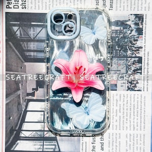 May include: Clear phone case with a pink lily and light blue butterflies. The case has a light blue border and the word "LUCKY" is printed on the bottom. The background is a newspaper print.