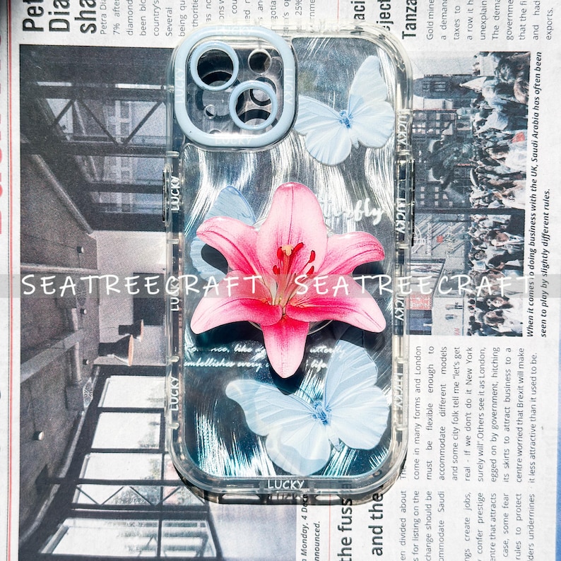 May include: Clear phone case with a pink lily and light blue butterflies. The case has a light blue border and the word "LUCKY" is printed on the bottom. The background is a newspaper print.