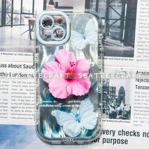 May include: Clear phone case with a floral and butterfly design. The case features a large pink hibiscus flower and two light blue butterflies. The words "LUCKY" and "SEATREECRAFT" are printed on the case.