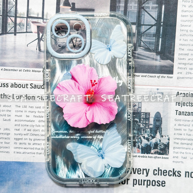 May include: Clear phone case with a floral and butterfly design. The case features a large pink hibiscus flower and two light blue butterflies. The words "LUCKY" and "SEATREECRAFT" are printed on the case.