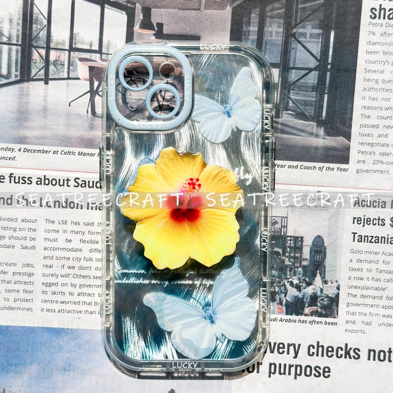 May include: Clear phone case with a light blue bumper. The case features a yellow hibiscus flower and two light blue butterflies. The words "CRAFT SEATREE" are printed on the case. The word "LUCKY" is printed on the case.