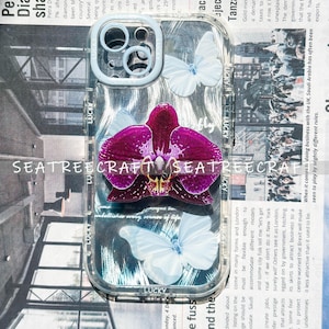 May include: Clear phone case with a purple orchid and white butterflies. The case has a light blue camera ring and the word "LUCKY" printed on the side. The background is a newspaper print.