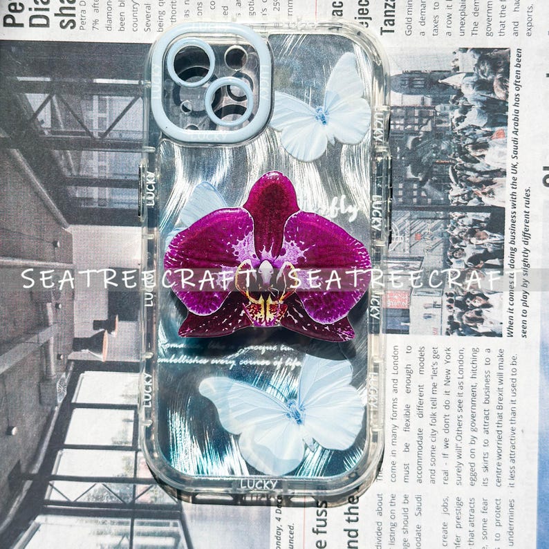 May include: Clear phone case with a purple orchid and white butterflies. The case has a light blue camera ring and the word "LUCKY" printed on the side. The background is a newspaper print.