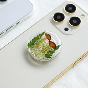 Puede incluir: Una agarradera de teléfono transparente con un diseño prensado de hongos y helechos. La agarradera es redonda y tiene un borde elevado.