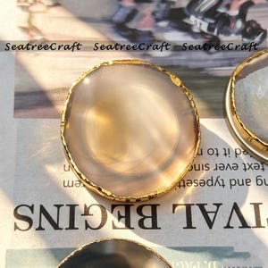 May include: A circular, translucent, light beige agate slice coaster featuring a polished surface and a textured, metallic gold-colored electroplated edge. The text SeatreeCraft is visible in the background, along with printed text.