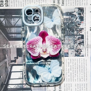 May include: Clear phone case with a purple and white orchid and white butterflies. The case has the word "LUCKY" printed on it. The case is designed to fit a phone.