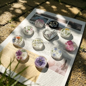 Peut inclure: Une collection de breloques circulaires en résine contenant des fleurs et de la verdure pressées, ainsi qu'une breloque en forme de cœur avec une lune, des étoiles et un petit cœur argentés. Le tout est disposé sur un journal avec le texte SeatreeCraft.