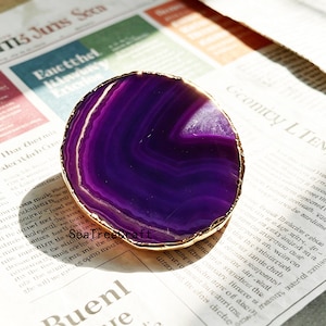 Peut inclure: Une tranche d'agate circulaire violette servant de dessous de verre, présentant des bandes concentriques de tons violet foncé et plus clair. La pierre a une surface polie et translucide, bordée d'une garniture dorée métallique aux contours irréguliers.