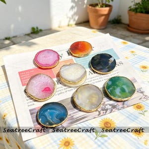 May include: A collection of eight circular agate coasters with polished surfaces and metallic gold-toned edges. The coasters feature various natural stone colors including pink, orange, white, black, green, and blue, arranged on a printed paper sheet.