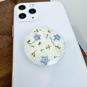 Puede incluir: Una agarradera de teléfono transparente con un fondo blanco y flores azules de nomeolvides prensadas en su interior.