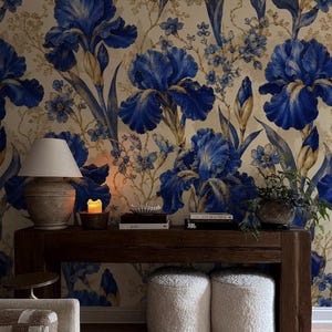 Puede incluir: Interior de salón con consola de madera oscura, lámpara, libros y vela. Dos pufs blancos y esponjosos debajo. El papel pintado presenta grandes flores de iris azules.