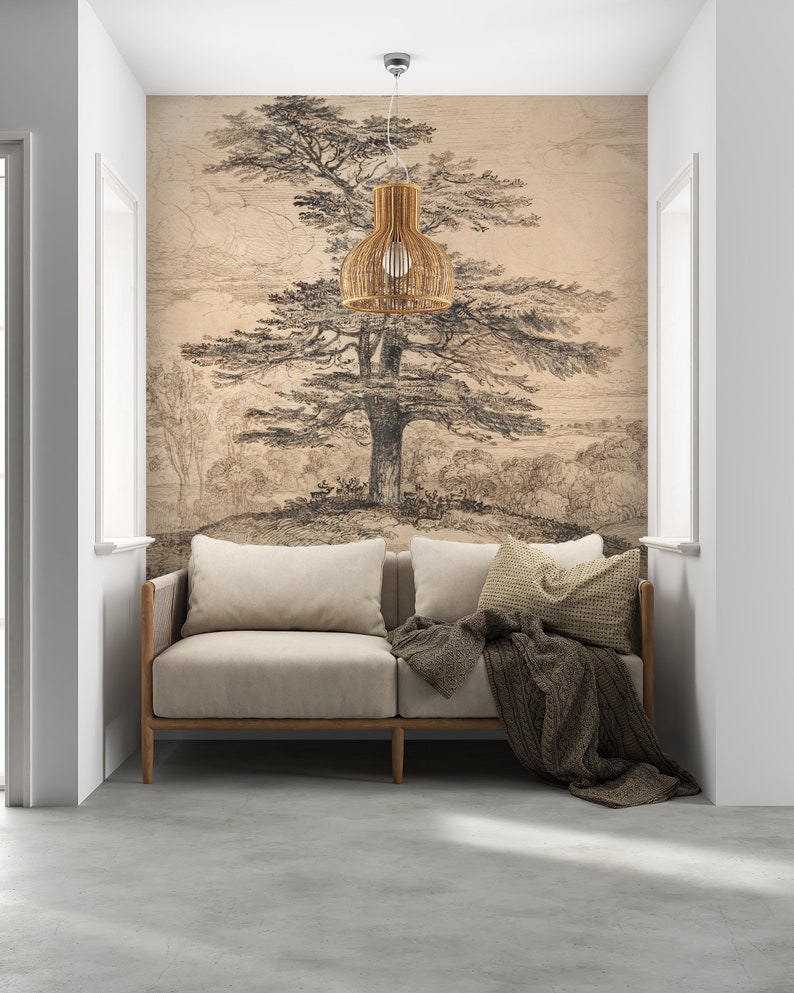 May include: A light beige sofa with a brown blanket and pillows sits in a room with a white wall and a large drawing of a tree on the wall. A woven pendant light hangs above the sofa.