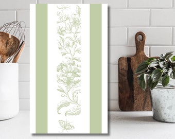 Puede incluir: Una toalla de cocina blanca con un diseño floral verde y rayas verdes a los lados. La toalla está colgada en una pared de azulejos blancos.