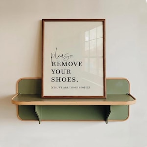 Op de afbeelding: Een ingelijst wandbord met een donkerhouten rand met de zwarte tekst: please REMOVE YOUR SHOES. (YES, WE ARE THOSE PEOPLE). Het bord staat op een horizontale olijfgroene metalen plank met afgeronde hoeken en een lichtbruine rand.