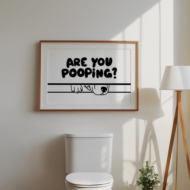 May include: A framed print with the text "ARE YOU POOPING?" in bold black letters. Below the text, a black line and a cartoon dog's face peeking over it. The print is in a wooden frame, above a white toilet.