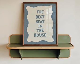 May include: A framed wall art print featuring the text THE BEST SEAT IN THE HOUSE in blue letters on a cream background with a wavy blue border, displayed on a green and light wood shelf with rounded edges and a dark wood frame.