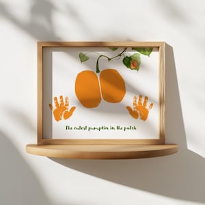 May include: A framed print featuring an orange pumpkin design with handprints, and the text "The cutest pumpkin in the patch." The artwork is displayed on a wooden shelf, perfect for fall decor.