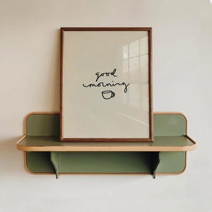 Può includere: Una stampa incorniciata con le parole "good morning" e un'illustrazione di una tazza di caffè. La cornice è marrone e la stampa è esposta su una mensola a muro verde e beige.