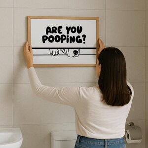 May include: A framed print with the text "ARE YOU POOPING?" in bold black letters. Below the text is a line with a dog's face peeking over. The print is held by a person in a bathroom setting.
