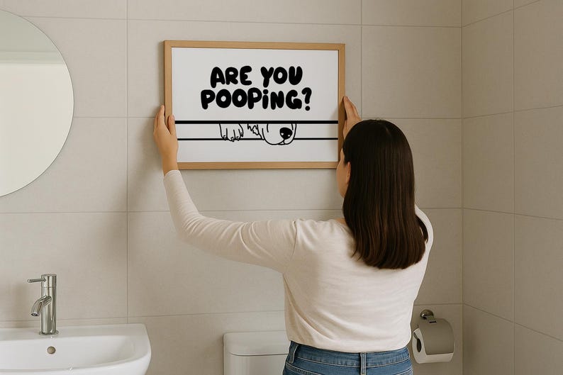 May include: A framed print with the text "ARE YOU POOPING?" in bold black letters. Below the text is a line with a dog's face peeking over. The print is held by a person in a bathroom setting.