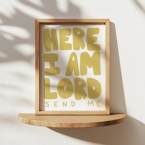 May include: A framed print with the words "HERE I AM LORD SEND ME" in a mustard yellow color. The print is in a light brown wooden frame and rests on a wooden shelf against a white wall.